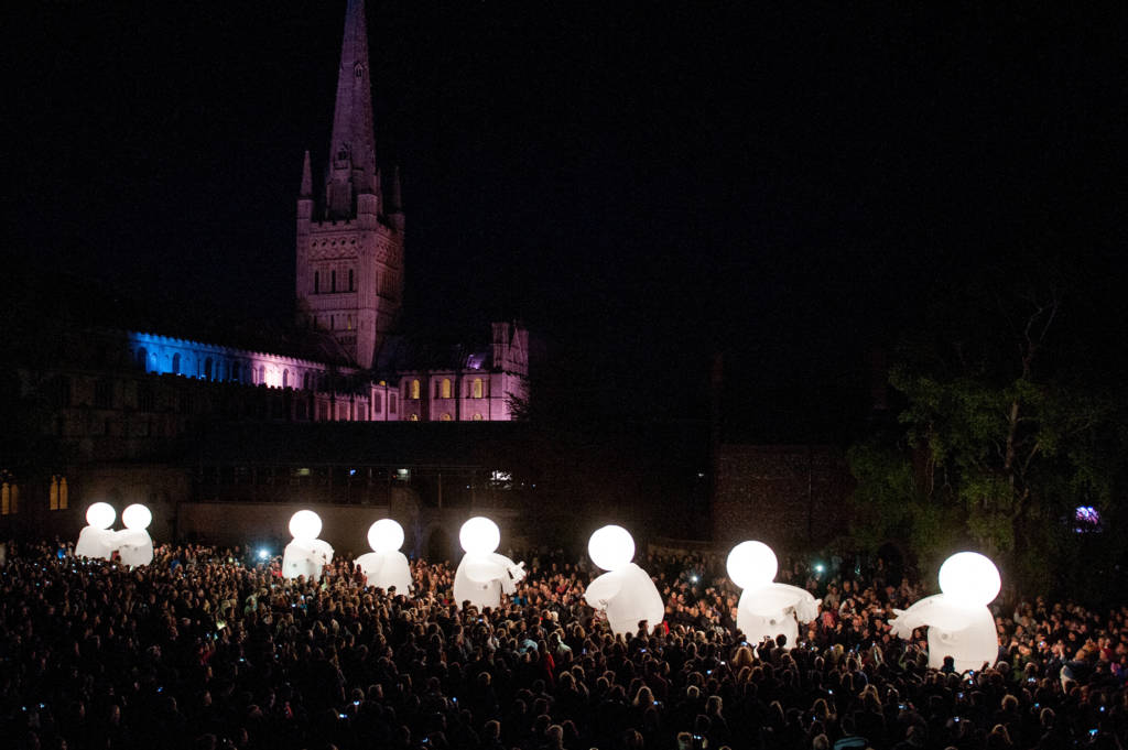 Hundreds are gathered outside Norwich Cathedral at night, observing a show of large illuminated inflatable puppets